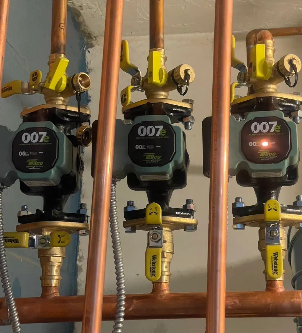 Close-up view of three green 007e circulation pumps attached to copper pipes with yellow on/off handles. The pumps are neatly aligned and connected to braided metal tubing.