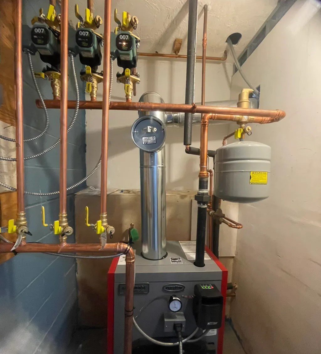A boiler installation featuring a red and gray unit with a silver exhaust pipe, connected to multiple copper pipes and green circulation pumps. The setup also includes a large expansion tank and electrical wiring in a compact utility space.