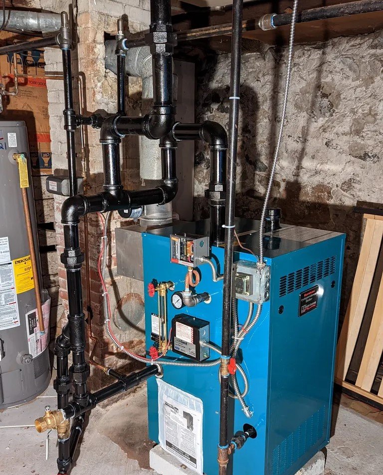A blue boiler system with black piping in a basement, surrounded by a brick wall and utility connections.