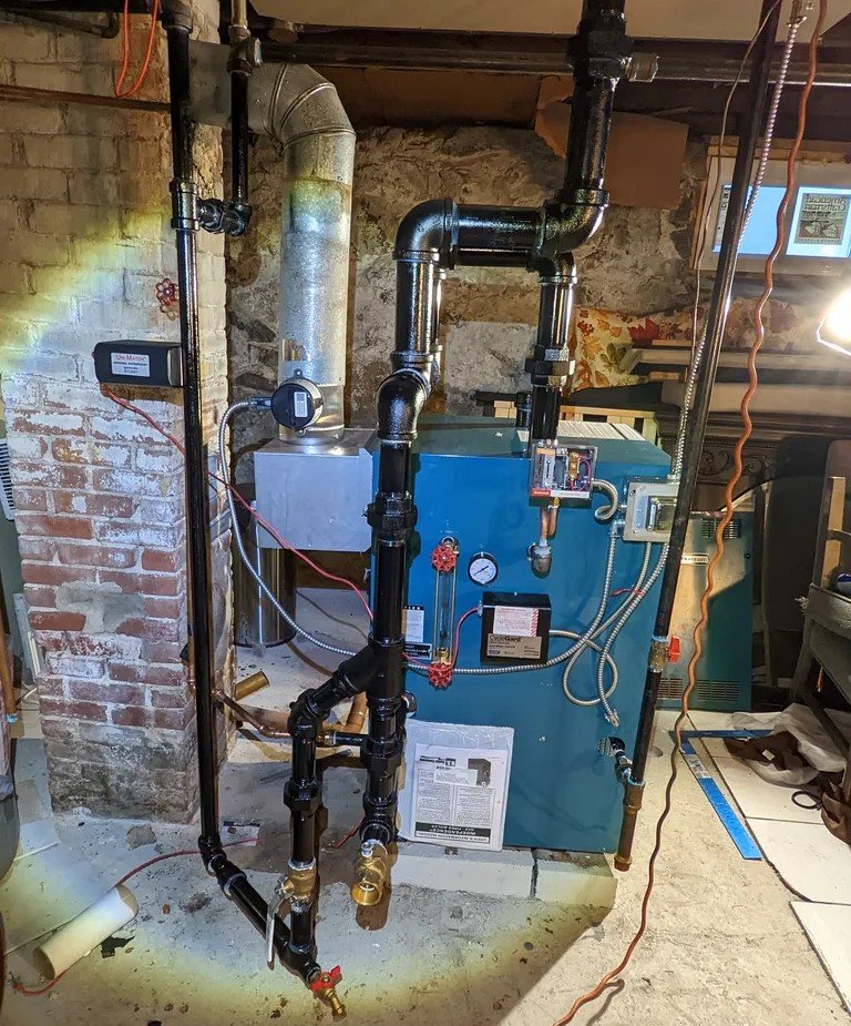 A basement installation featuring a blue boiler unit connected to black iron pipes. The system includes a pressure gauge, manual valves, and a visible exhaust vent leading to the ceiling.