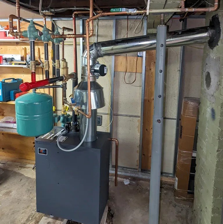 A residential boiler setup with copper piping, a green expansion tank, and a stainless steel vent pipe in a workshop area.