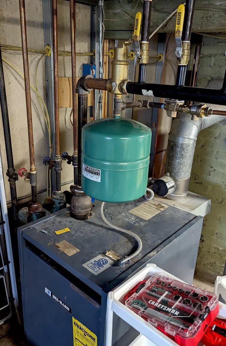 A close-up of the green expansion tank and copper pipe connections above a boiler unit. The setup includes multiple shut-off valves with yellow handles and a silver flex connector.