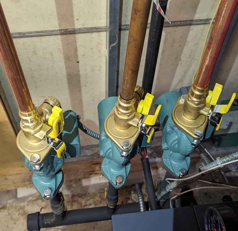 A close-up of three green circulation pumps attached to copper pipes. The pumps have bright yellow valve handles and are securely mounted in a neatly organized configuration.