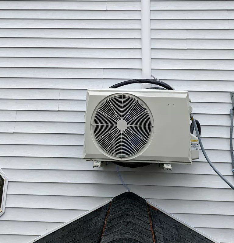 Close-up of an outdoor air conditioning unit mounted on the exterior wall of a house with white siding, positioned above a roofline.