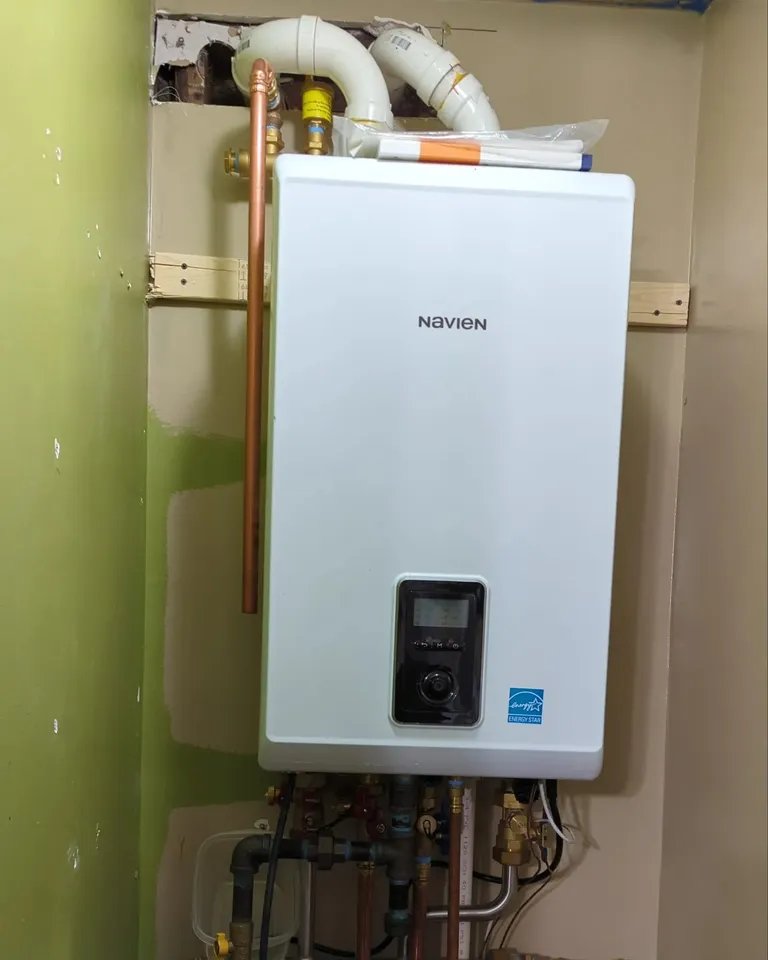Close-up of a Navien tankless water heater mounted on a green wall, featuring copper pipe connections and energy-efficient labeling.